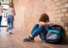 Sad child sitting against brick wall, kids running away.