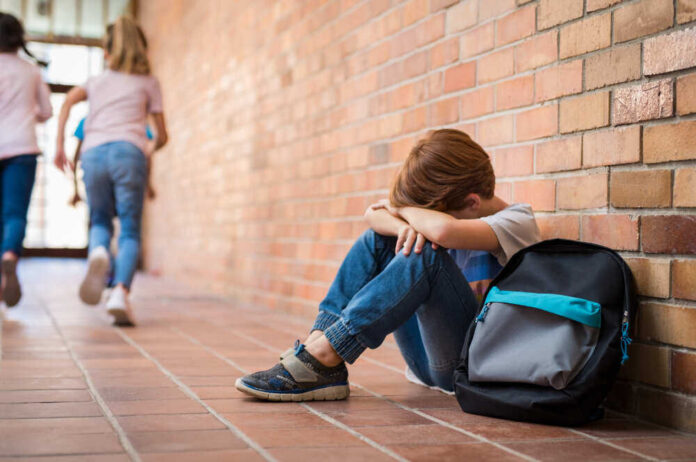 Sad child sitting against brick wall, kids running away.