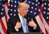 Donald Trump speaking in front of multiple American flags.