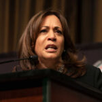 A woman speaking at a podium, looking serious.