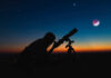 Silhouette of person using telescope against colorful twilight sky.