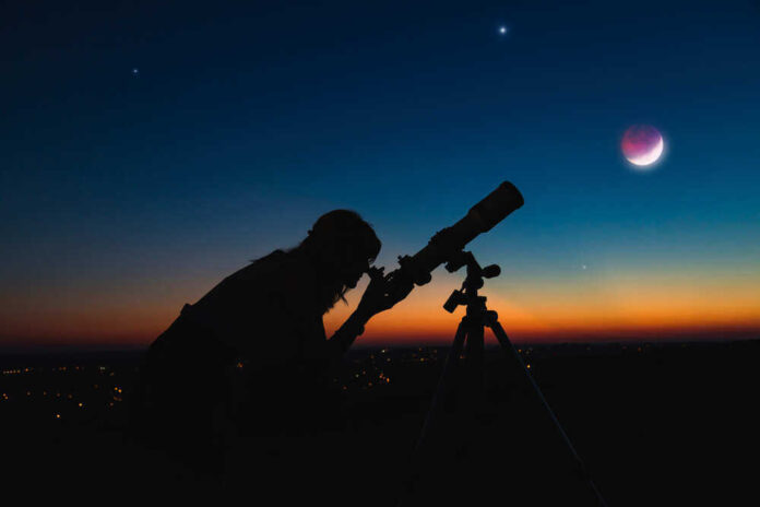 Silhouette of person using telescope against colorful twilight sky.