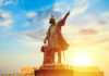 Bronze statue pointing skyward with sunlight background.