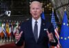 Joe Biden in suit speaking, international flags in background.