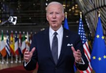 Joe Biden in suit speaking, international flags in background.