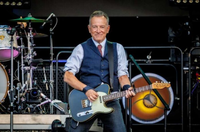 Bruce Springsteen playing electric guitar on stage, drum set behind.