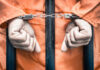 Handcuffed person in orange jumpsuit behind prison bars.