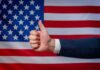 Thumbs up in front of American flag background.