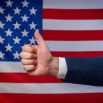 Thumbs up in front of American flag background.