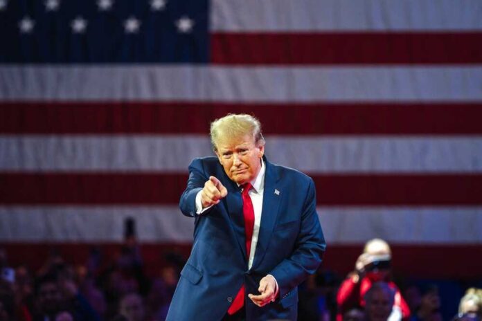 Donald Trump in front of American flag pointing toward camera.