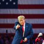 Donald Trump in front of American flag pointing toward camera.