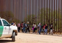 People walking along a border fence with patrol vehicle.