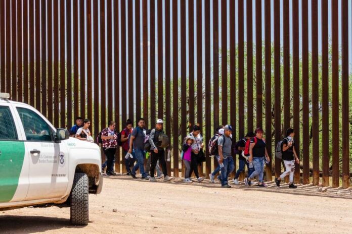 People walking along a border fence with patrol vehicle.