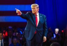 Donald Trump in suit pointing on stage with audience behind.