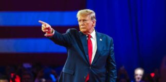 Donald Trump in suit pointing on stage with audience behind.