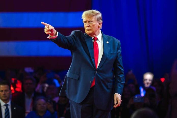 Donald Trump in suit pointing on stage with audience behind.