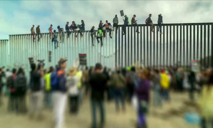 People sitting on a border wall with crowd below.