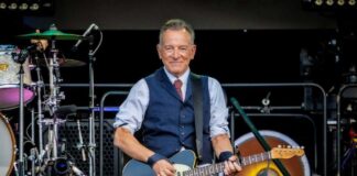 Bruce Springsteen playing electric guitar on stage, drum set behind.