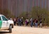 People walking along a border fence with patrol vehicle.