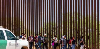 People walking along a border fence with patrol vehicle.