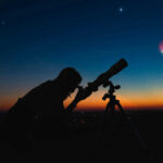 Silhouette of person using telescope against colorful twilight sky