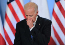 Joe Biden coughing in a suit standing in front of flags