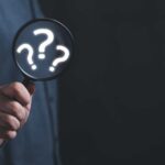 SPIRITUAL SCAM? Michigan AG Targets “Twin Flames” Cult (Video) Hand holding magnifying glass with question marks inside.