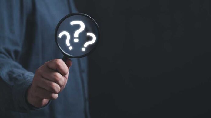 Hand holding magnifying glass with question marks inside.