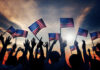 Silhouetted crowd holding American flags against a sunset