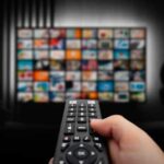 Hand holding a remote control in front of a television displaying various media options