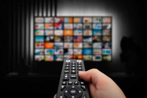 Hand holding a remote control in front of a television displaying various media options