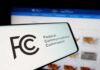 Close-up of a mobile device displaying the FCC logo with a website in the background