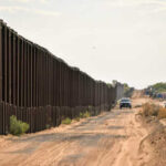 A long border wall stretching through a desert landscape with vehicles nearby