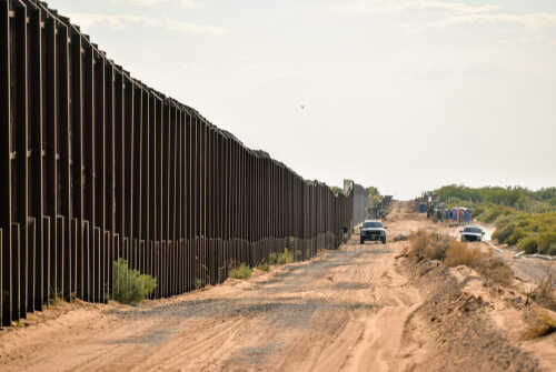 A long border wall stretching through a desert landscape with vehicles nearby
