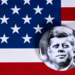 A black and white badge of John F. Kennedy on an American flag background