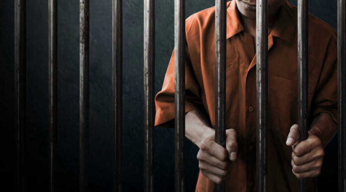 Man in orange prison uniform behind jail bars.