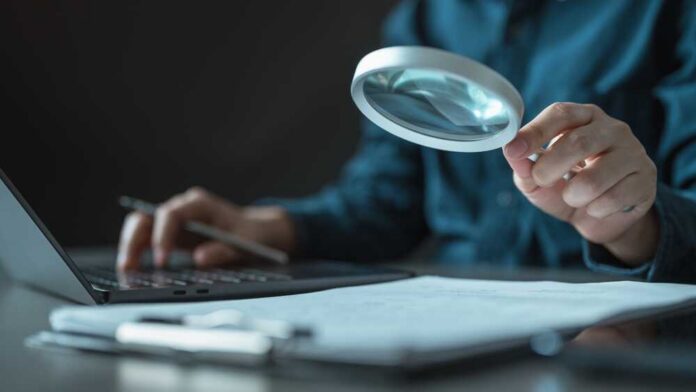 shutterstock_2708642557.jpg Person using a magnifying glass while working on a laptop