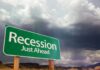 A green road sign indicating 'Recession Just Ahead' against a cloudy sky