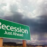 A green road sign indicating 'Recession Just Ahead' against a cloudy sky