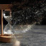 Hourglass disintegrating into specks and particles, dark background.