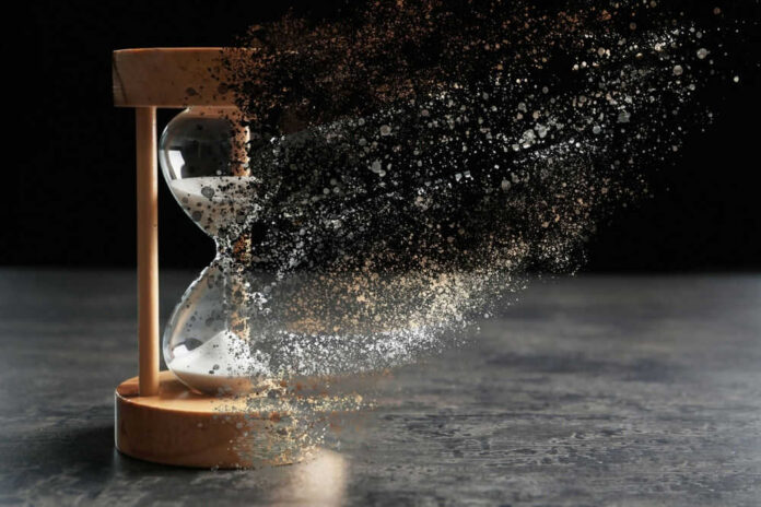 Hourglass disintegrating into specks and particles, dark background.
