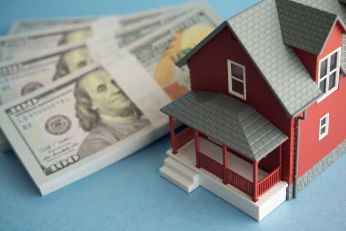 shutterstock_2600980667.jpg A model red house next to stacks of hundred-dollar bills