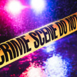 Crime scene tape with flashing lights in background.