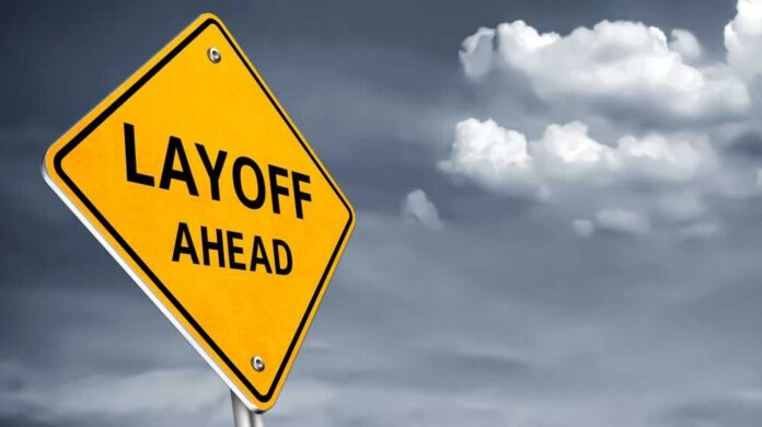 shutterstock_2503823051.jpg Yellow warning sign indicating layoffs ahead against a cloudy background