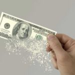Army Budget Hole Triggers Training Axe Hand holding a disintegrating hundred dollar bill.