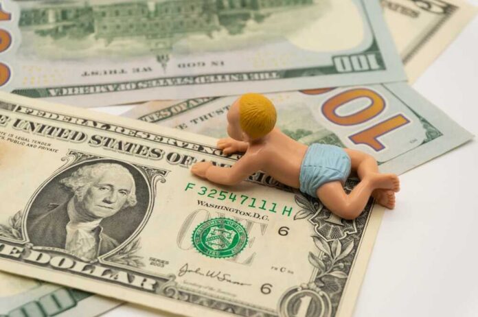 Miniature,Baby,Toys,On,The,Table,With,Dollar,Bills.,A A baby figurine crawling on top of various dollar bills