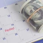 A rolled bundle of cash placed on a calendar marked with 'Payday!'