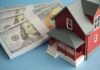 A model red house next to stacks of hundred-dollar bills