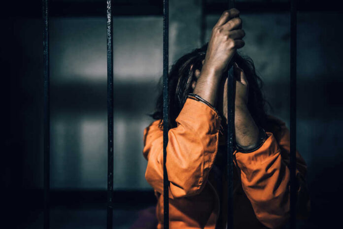 Person in orange prison uniform behind bars, head down.