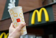 Fast-Food Arms Race: McDonald’s Goes ALL-IN Hand holding McDonalds cup in front of restaurant sign.